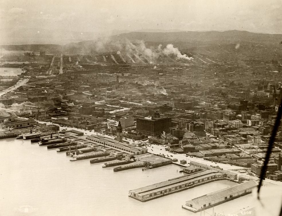 Aerial photograph of San Francisco's Embarcadero waterfront in the 1920s. The Ferry Building clock tower stands at center; finger piers extend into the Bay along the bottom of the frame; the urban core stretches from waterfront to hills, with industrial smokestacks producing visible smoke across the city skyline.