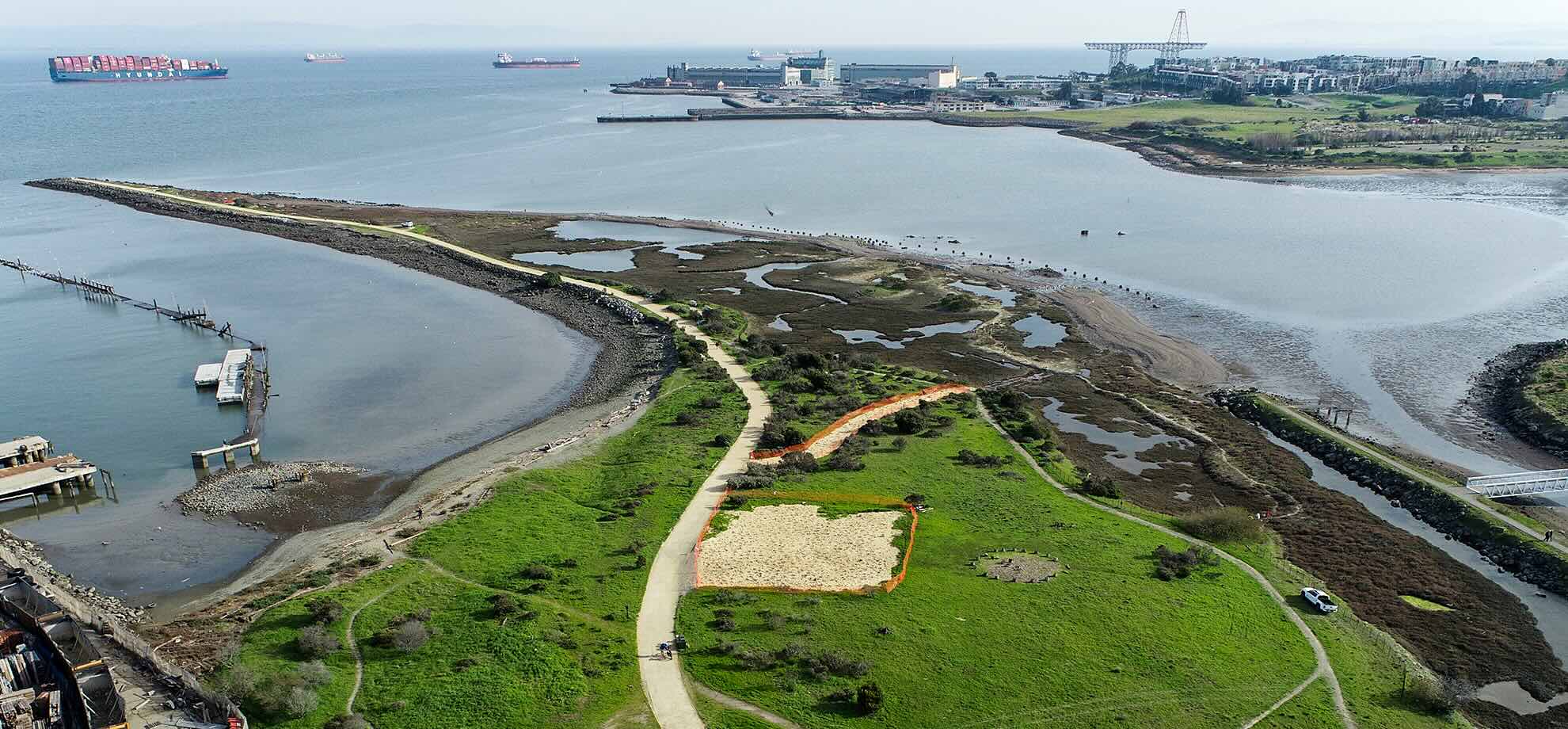 Aerial photograph of Heron's Head Park, San Francisco, looking east-southeast. The restored wetland tidal channels are visible in the foreground with an orange-bordered restoration plot; in the background across the channel, the former Hunters Point Naval Shipyard with its iconic gantry crane, the new San Francisco Shipyard residential development on Hunters Point Hill, and container ships and tankers in the bay.
