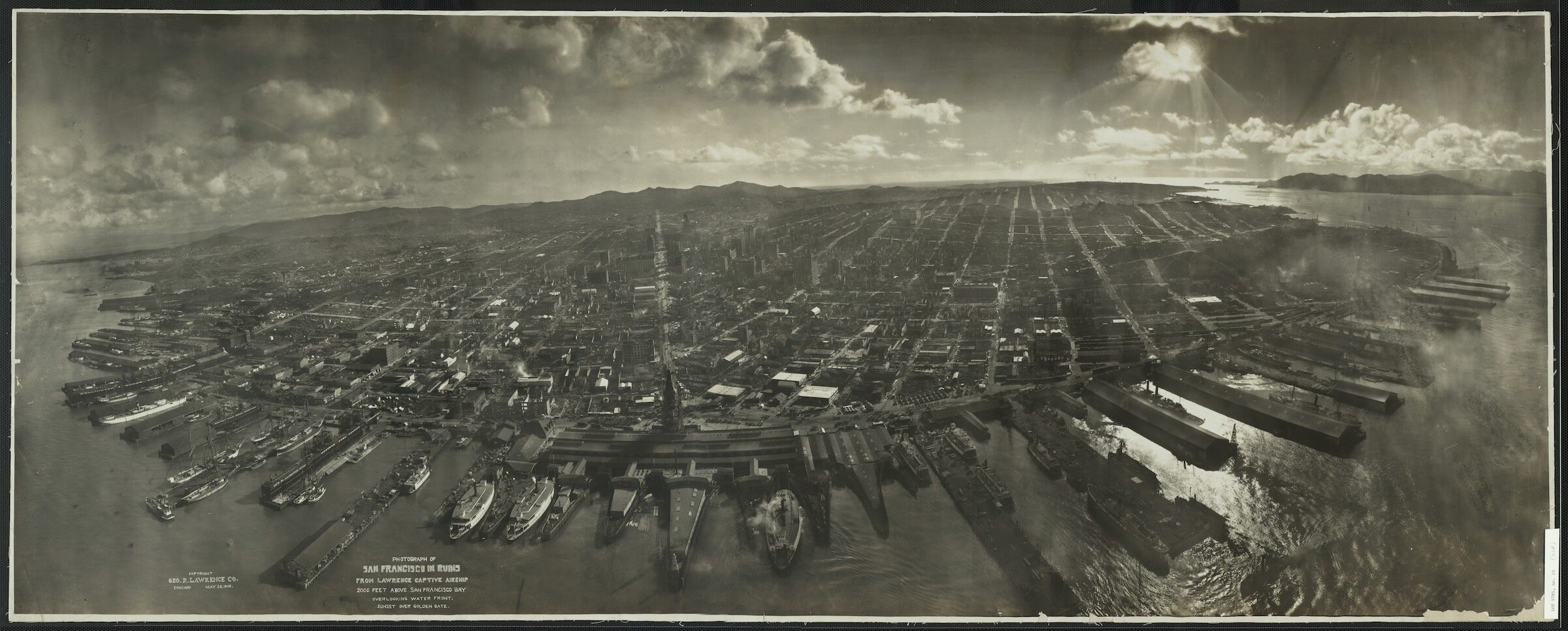 George R. Lawrence panoramic photograph titled 'San Francisco in Ruins,' taken in 1906 from a captive airship roughly 2,000 feet above the Bay. The image shows the working waterfront and its finger piers intact along the bay, with the city stretching inland; smoke rises from the burned downtown five weeks after the April 1906 earthquake and fire.